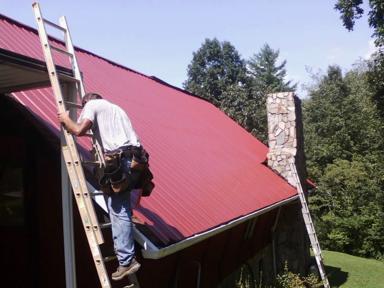 Roofer on a ladder beside a red metal roof and stone chimney — Soffit & Fascia Repair by Roofing Brothers in Ola